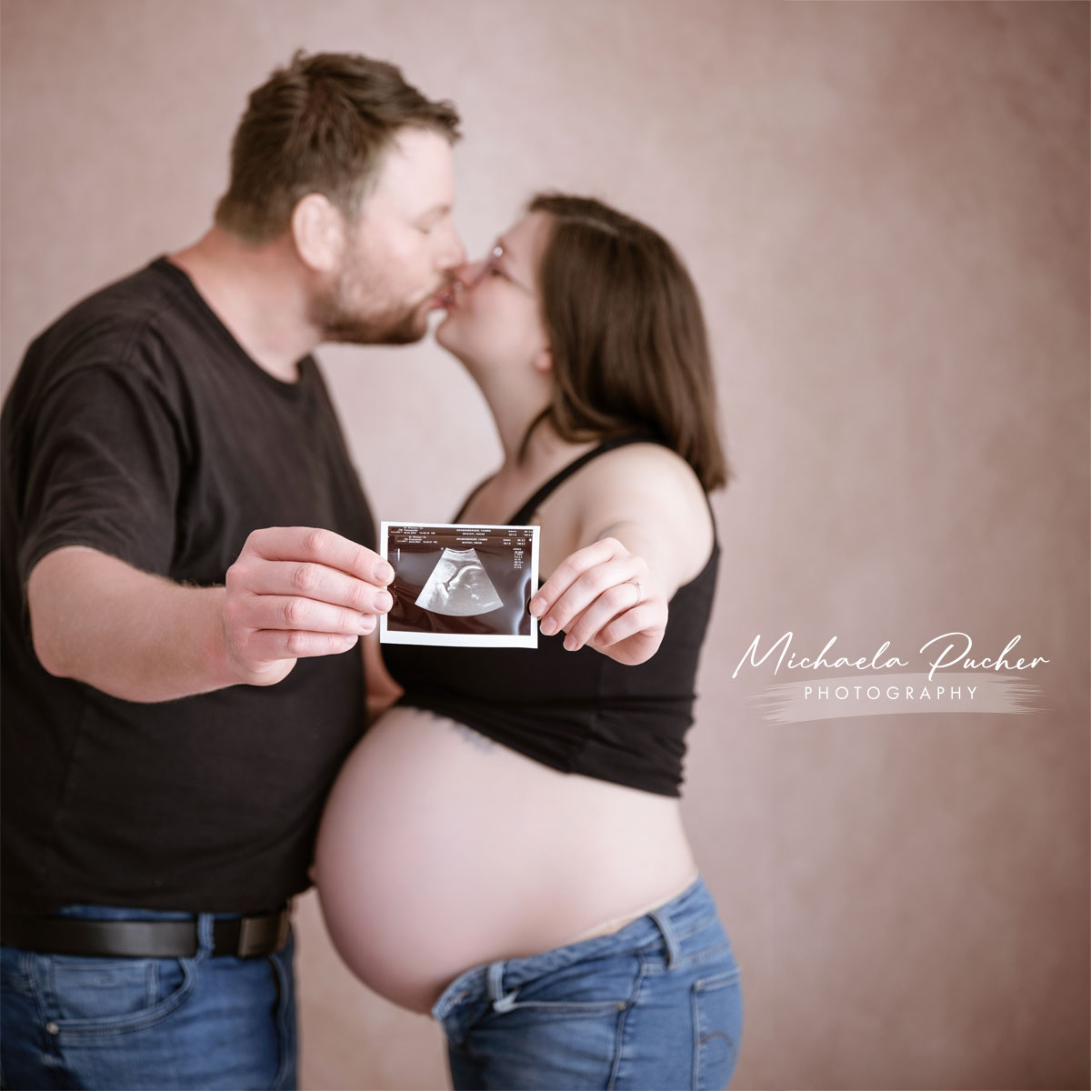 Schwangere Frau und ihr Partner küssen sich, während sie gemeinsam ein Ultraschallbild ihres Babys in die Kamera halten.